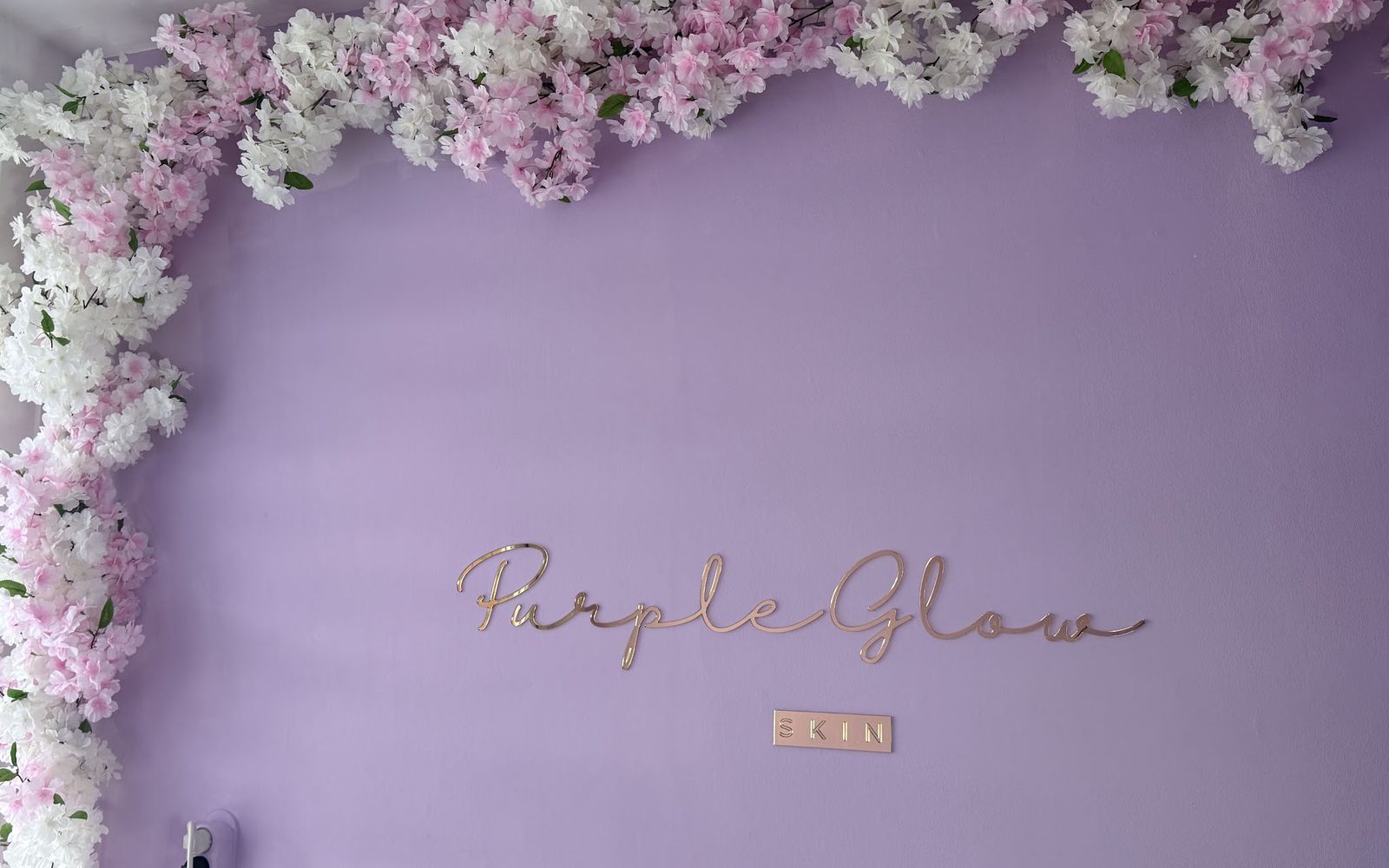 De lila salonwand van PurpleGlow Skin met een slinger van roze en witte bloesem en de rosé-gouden letters PurpleGlow Skin