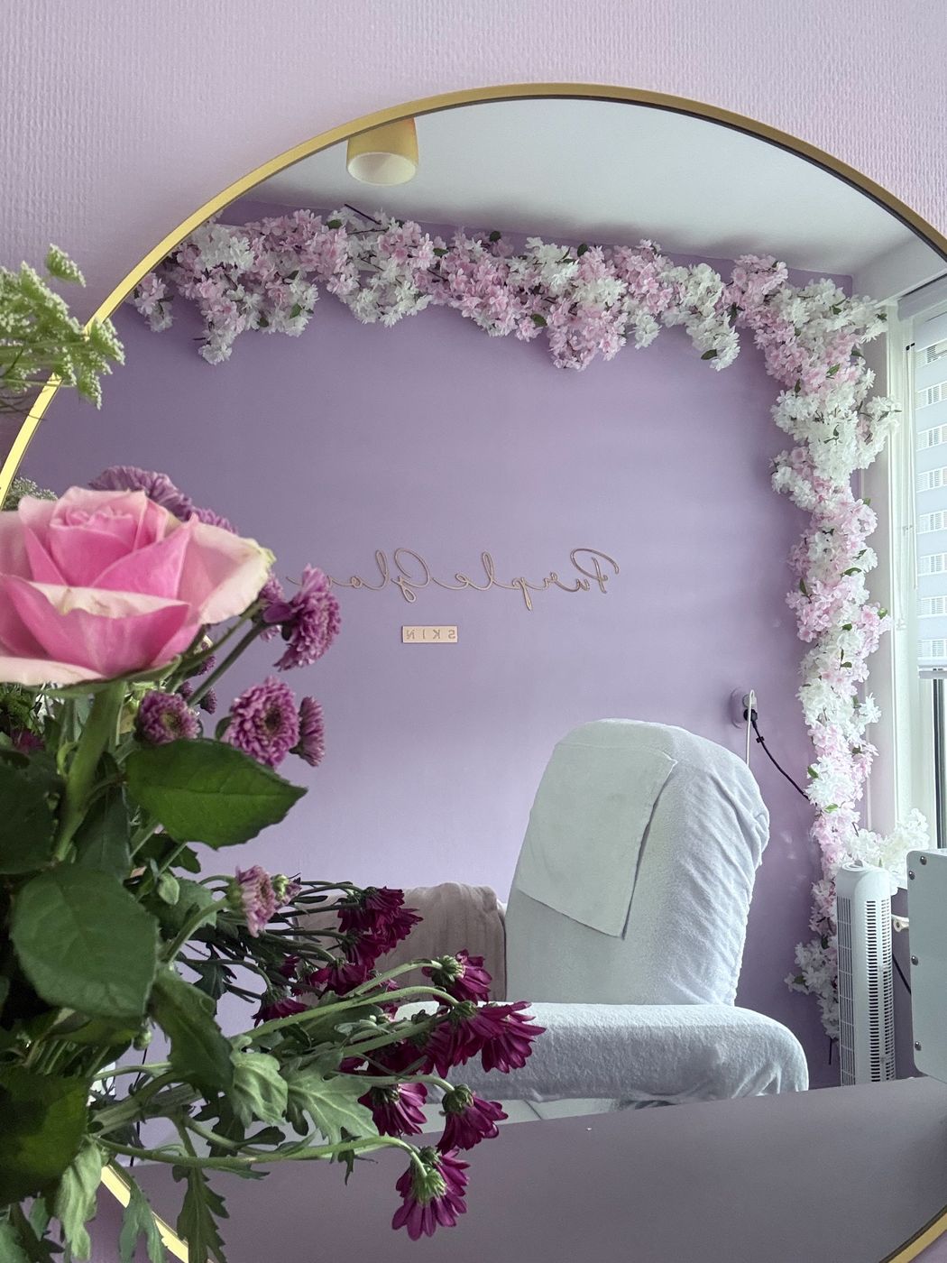 Roze rozen voor de ronde spiegel met gouden rand, waarin de behandelstoel en de bloesemwand te zien zijn